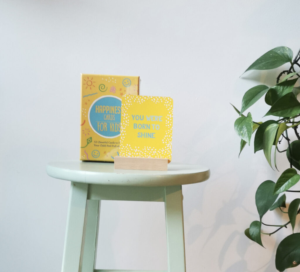 Yellow box of Happiness cards on a green stool with a green plant to the right